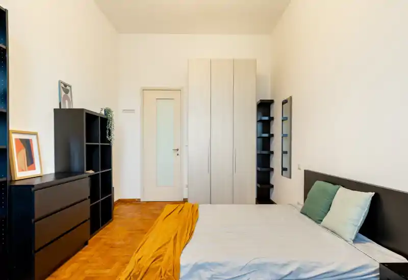 Camera da letto luminosa e ben arredata con letto matrimoniale, armadio e scaffalature; pavimento in legno e tessili caldi rendono l'ambiente accogliente e adatto per annunci.