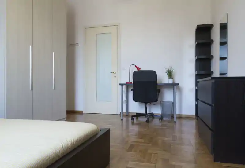 Camera da letto luminosa e minimale con letto ben visibile in primo piano, armadio, scrivania e scaffali — pavimento in parquet e arredo neutro e ordinato.
