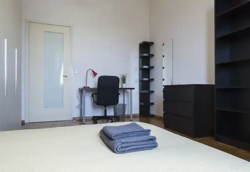 Camera da letto luminosa e minimal con un letto rifatto in primo piano, scrivania e scaffalature sullo sfondo e mobili contenitori scuri dal design moderno.