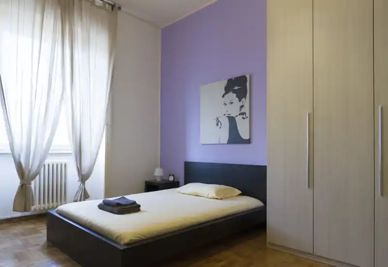 Camera da letto singola moderna e luminosa con letto basso in legno scuro, biancheria chiara, comodino e grande armadio. La luce naturale filtrata dalle tende alte crea un'atmosfera calma e accogliente.