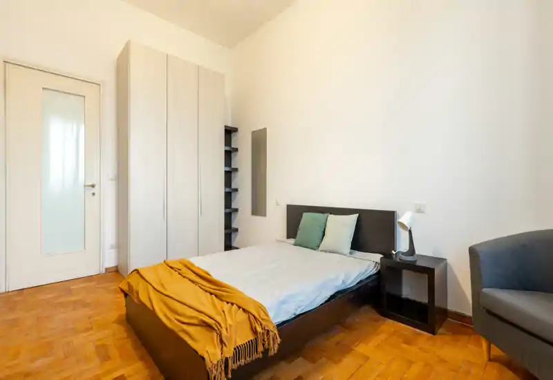Camera da letto luminosa e dal gusto semplice con letto matrimoniale, comodino e armadio; pavimento in legno e tessili caldi creano un ambiente accogliente.