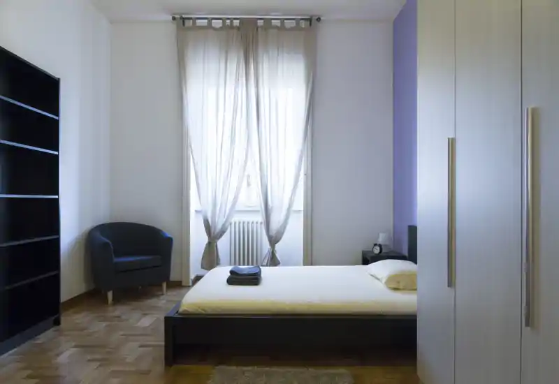 Camera da letto luminosa e minimal con letto matrimoniale, tende leggere e armadio. La stanza è ben arredata con luce naturale, una poltroncina compatta e pavimento in parquet.
