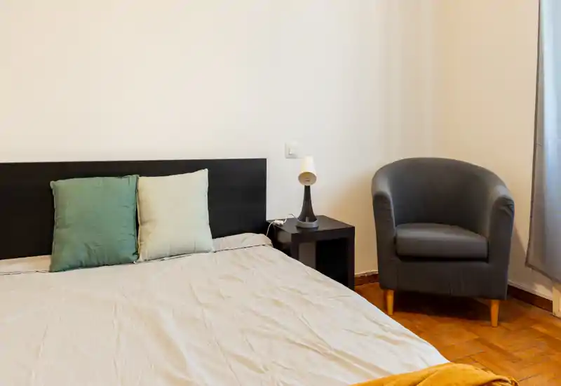 Camera da letto luminosa e minimale con un grande letto, due cuscini, comodino e una poltrona vicino alla finestra. Linee pulite e luce naturale morbida creano un'atmosfera calma e accogliente.