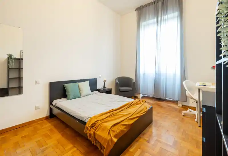 Camera luminosa e accogliente con letto matrimoniale, plaid e cuscini, poltroncina e scrivania vicino alla finestra—ideale per soggiorni rilassanti o affitti brevi.