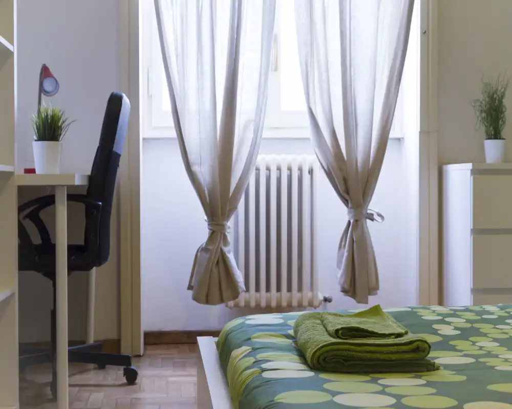 Camera da letto luminosa e ordinata con letto matrimoniale in primo piano e luce naturale dalla finestra; scrivania e sedia laterali la rendono adatta sia al relax che allo studio.