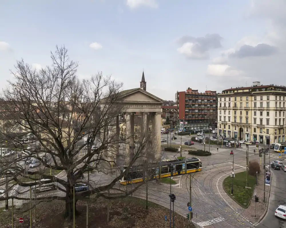 Veduta di una piazza urbana con un tram davanti a un edificio classico con colonne e palazzi circostanti; adatta come immagine esterna/stradale per un alloggio nelle vicinanze.