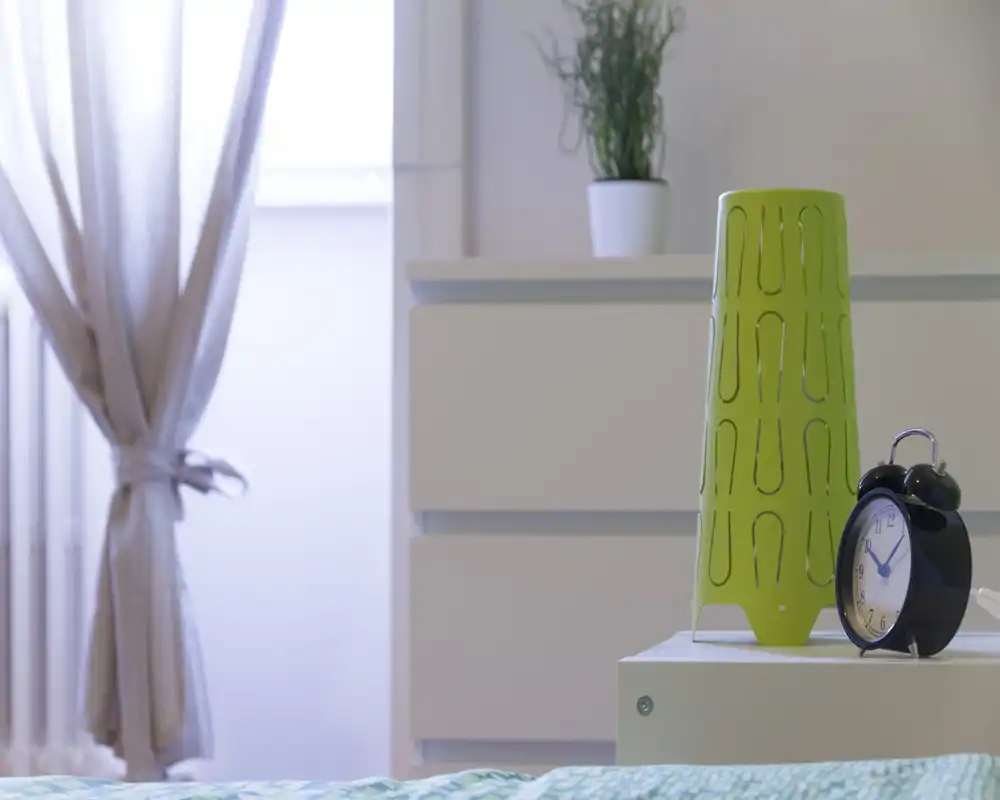 Angolo di camera luminoso e moderno con comodino, lampada decorativa e sveglia; luce naturale morbida dalla finestra con tende e una pianta sul comò per un’atmosfera accogliente.