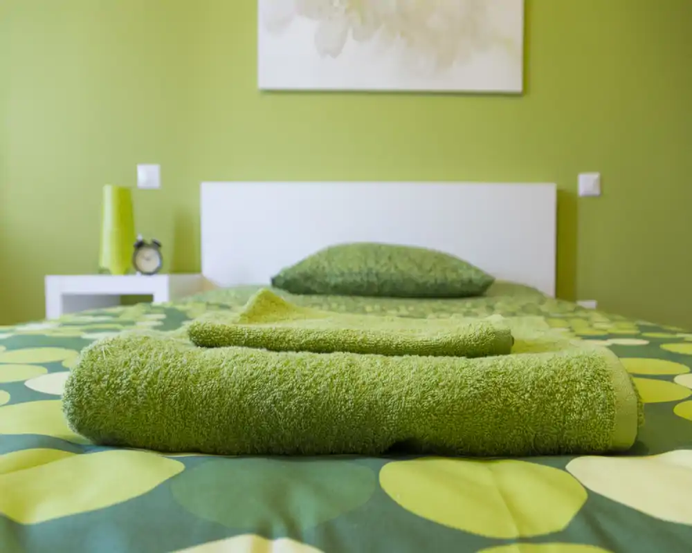 Camera da letto luminosa e ordinata con una palette verde; biancheria ben stirata e asciugamani piegati sul letto creano un allestimento accogliente.