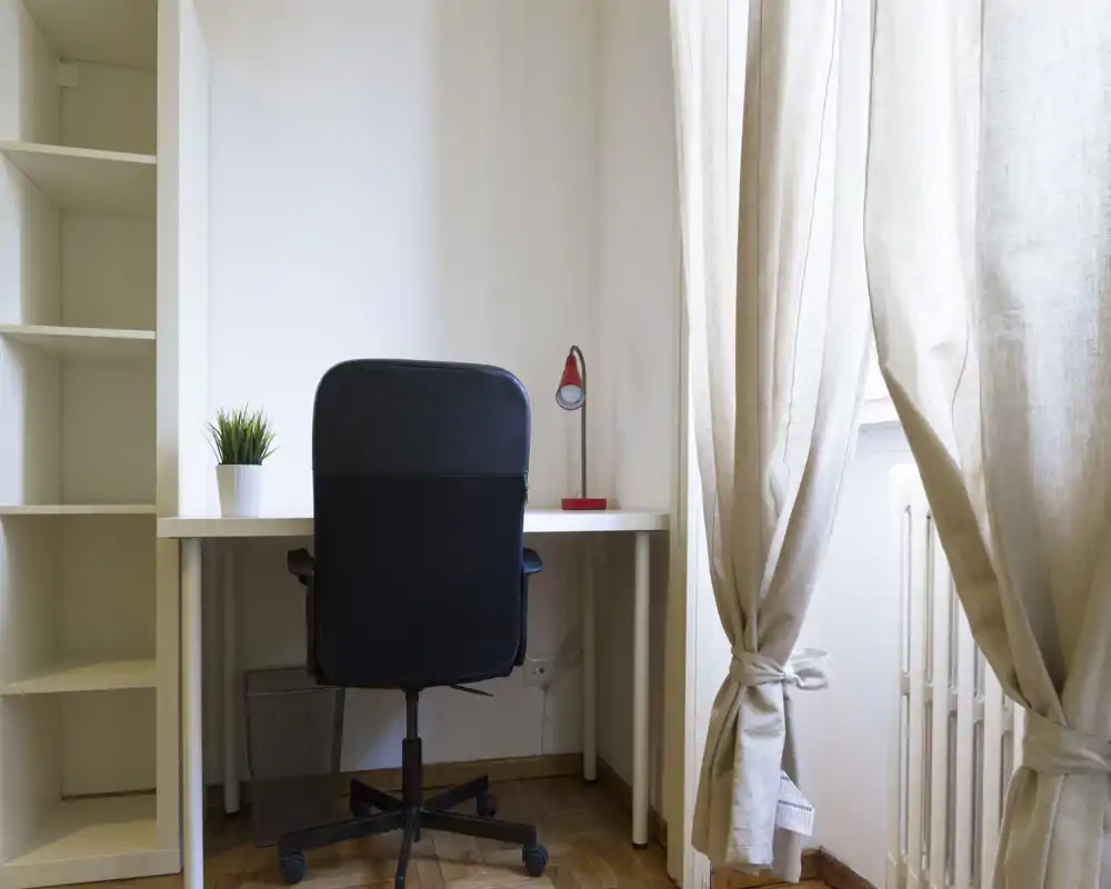 Angolo studio luminoso e minimale con scrivania bianca, sedia girevole nera, piccola lampada da tavolo rossa e una piantina vicino a una finestra con tende.