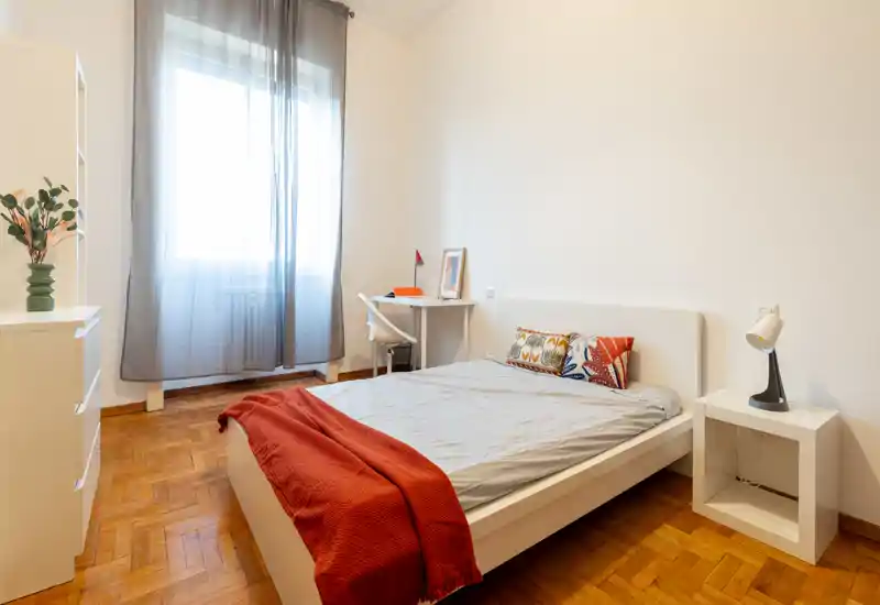 Camera da letto luminosa e minimal con letto matrimoniale, cuscini decorativi e coperta rossa; luce naturale da una finestra grande e una piccola scrivania nell'angolo.