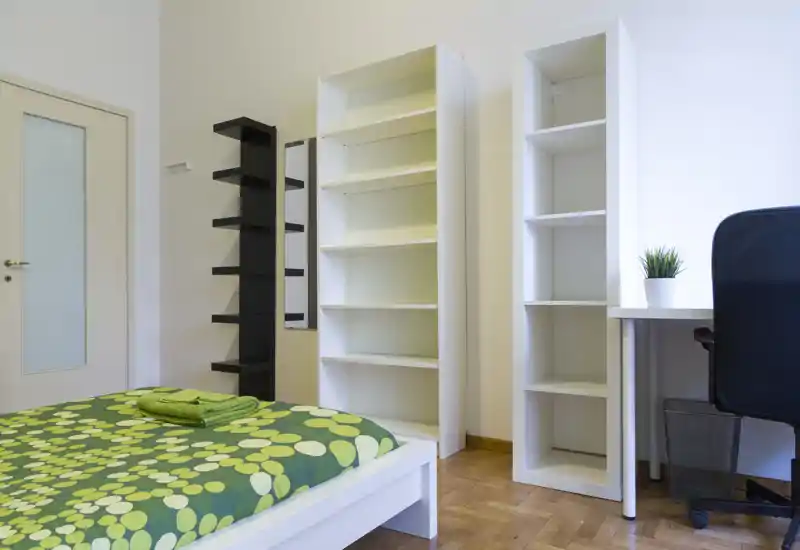 Camera da letto luminosa e minimal con struttura letto bianca, biancheria verde a fantasia e scaffali aperti; scrivania e sedia sulla destra per un layout funzionale.