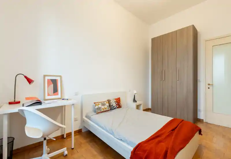 Camera singola ordinata e luminosa con letto bianco, cuscino decorativo e coperta rossa; armadio e scrivania compatta con sedia, ideale per studenti o single.