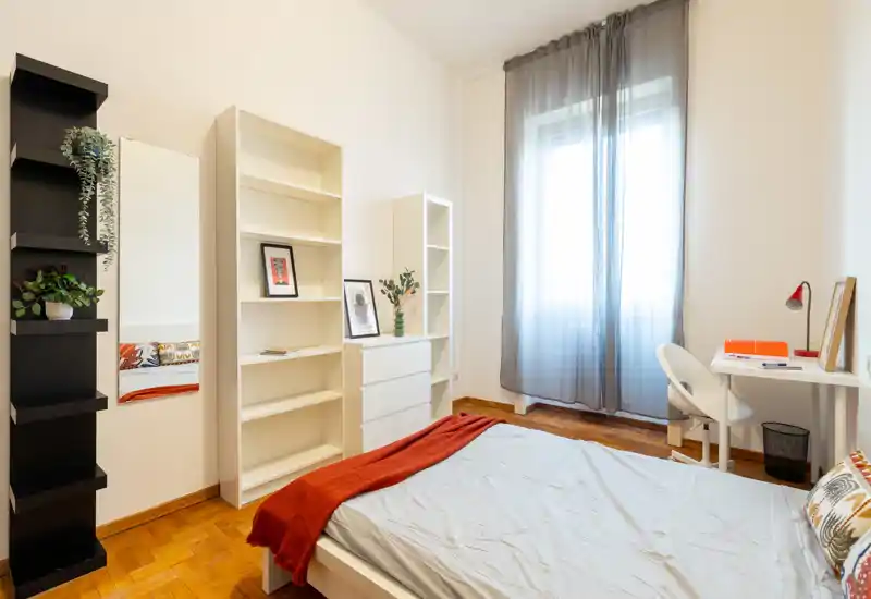 Camera da letto luminosa e minimal con letto matrimoniale, pavimento in legno, scrivania vicino alla finestra e scaffalature bianche con un tocco di colore rosso.