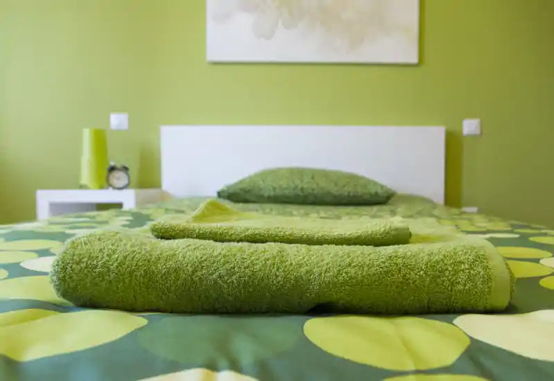Camera da letto luminosa e ordinata con una palette verde; biancheria ben stirata e asciugamani piegati sul letto creano un allestimento accogliente.