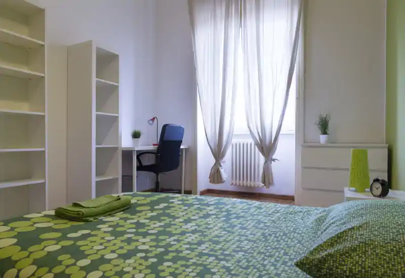 Camera luminosa e ben arredata con copriletto verde a pois, scrivania e scaffalature. La luce naturale dalla finestra crea un’atmosfera fresca e accogliente.