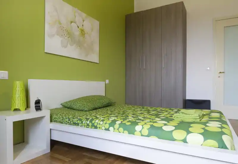 Camera da letto luminosa e moderna con letto matrimoniale con biancheria verde a motivo, comodino e armadio alto; arredo minimal e pavimento in legno caldo.