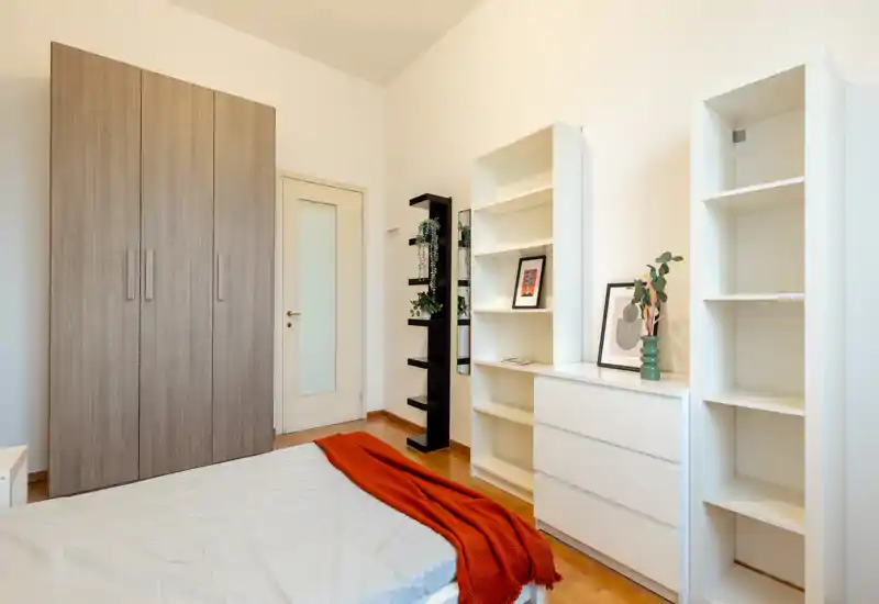 Camera da letto luminosa e ben allestita con letto visibile, scaffalature bianche e armadio — perfetta per annunci che valorizzano lo spazio e la luce naturale.