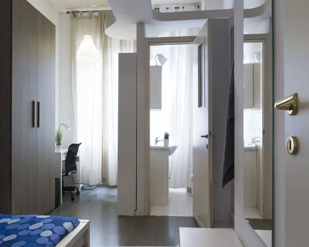 Camera da letto moderna e luminosa con letto parzialmente visibile in primo piano, ampia finestra con tende leggere, scrivania semplice e armadio — ambiente pulito e arioso ideale per affitti brevi.