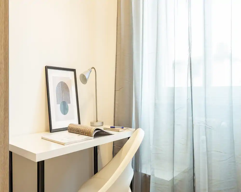 Un angolo studio luminoso e minimal con scrivania bianca, lampada moderna, quadro incorniciato e una sedia comoda accanto a tende leggere che filtrano la luce naturale.