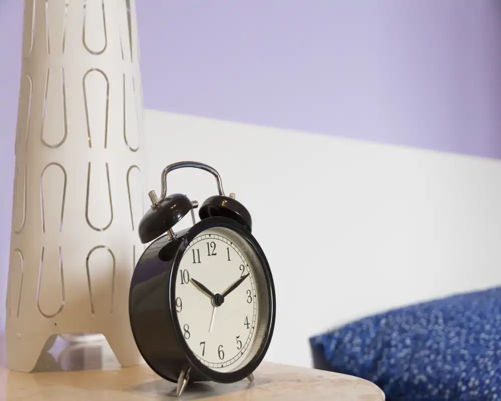 Primo piano di un comodino con una sveglia nera classica, una lampada decorativa e un cuscino blu visibile sul bordo: perfetto per mostrare i dettagli della camera e l'atmosfera mattutina.