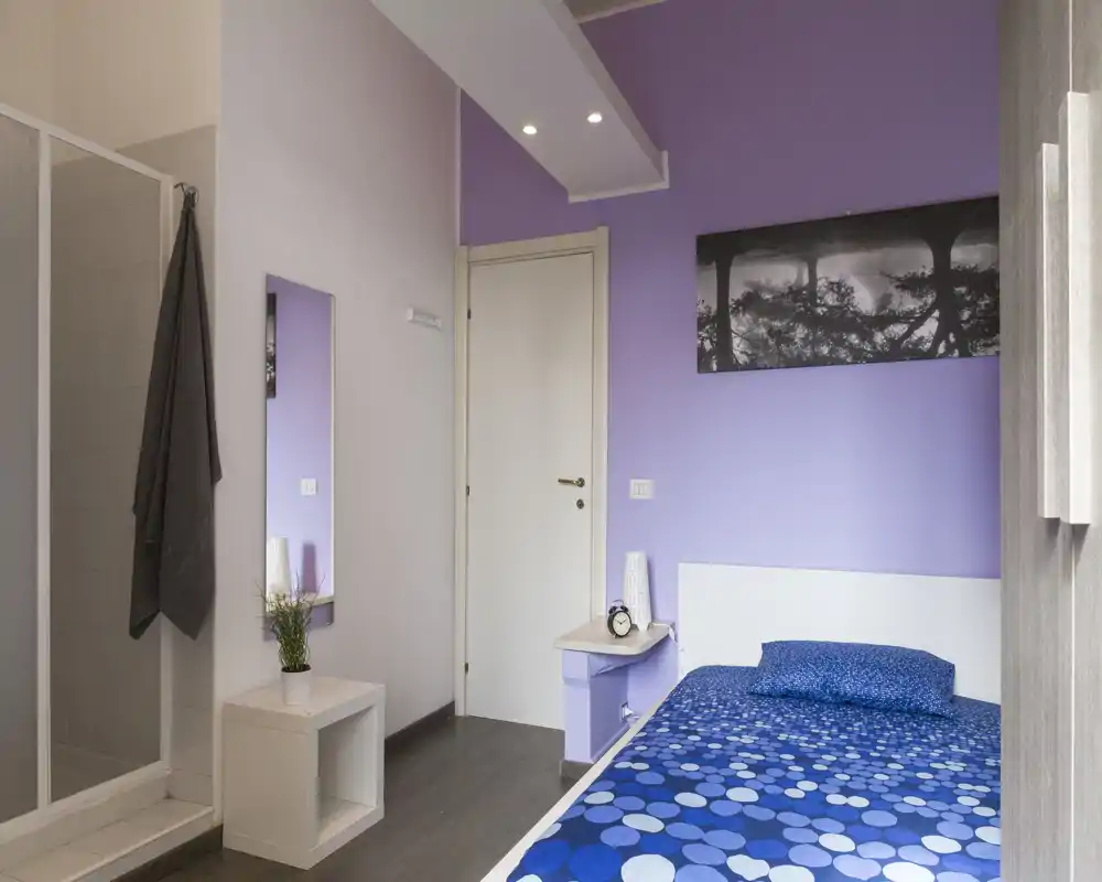 Camera singola ordinata con letto blu a fantasia, comodino e pareti lilla. La stanza è luminosa e ben arredata, adatta per annunci in affitto.
