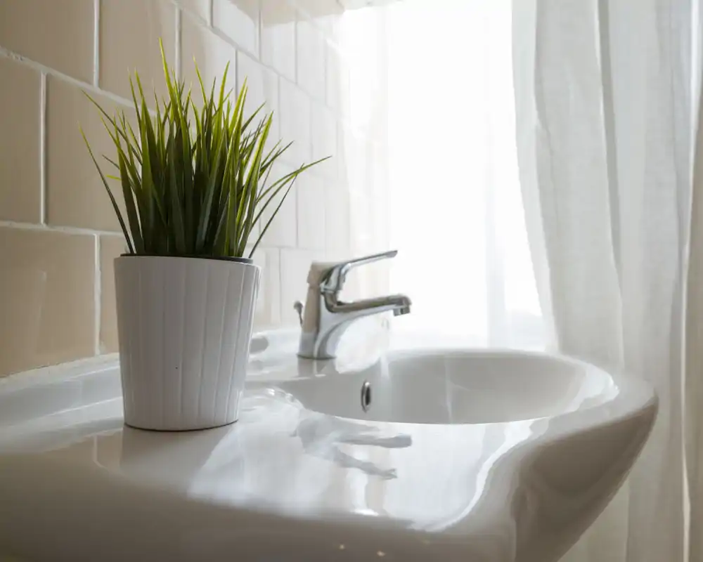 Lavabo da bagno moderno e luminoso con rubinetto cromato e una piccola pianta in vaso sul piano, su pareti piastrellate chiare e tende leggere che diffondono la luce naturale.