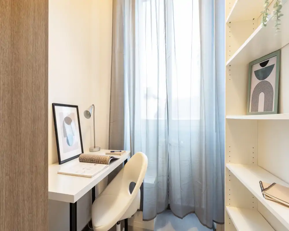 Angolo lavoro compatto e ben illuminato con scrivania bianca, sedia moderna, lampada da tavolo e scaffalatura — ideale per smart working o studio.