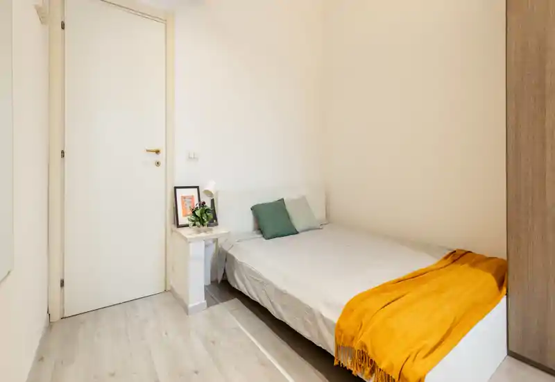 Habitación pequeña y luminosa con cama doble bien hecha, cojines verdes y una manta naranja. Paredes neutras y suelo claro que crean un ambiente moderno y tranquilo, perfecto para alquileres de corta estancia.