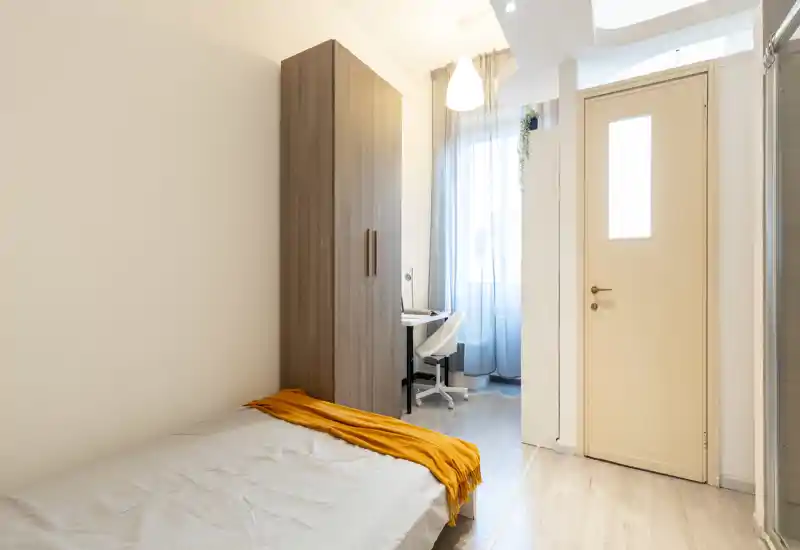 Dormitorio compacto y luminoso con cama individual, armario alto y un pequeño escritorio junto a la ventana. La luz natural y la manta mostaza crean sensación acogedora.