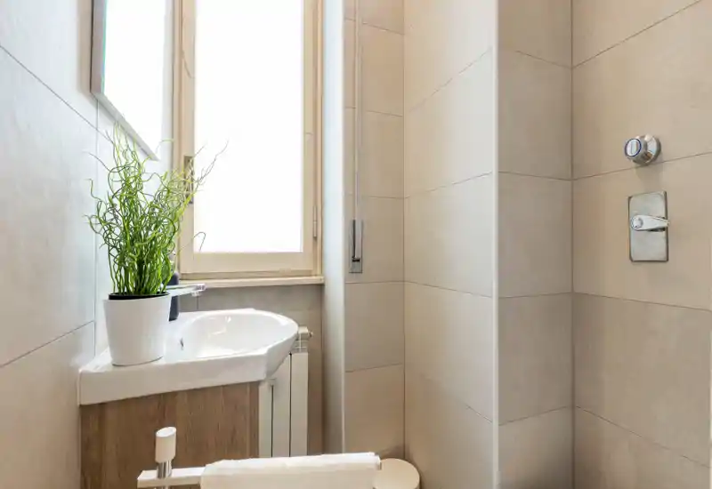 Bagno compatto e moderno con piccolo lavabo, comandi della doccia e pareti piastrellate chiaro. La luce naturale da una finestra satinata illumina l'ambiente e una pianta in vaso aggiunge un tocco fresco.