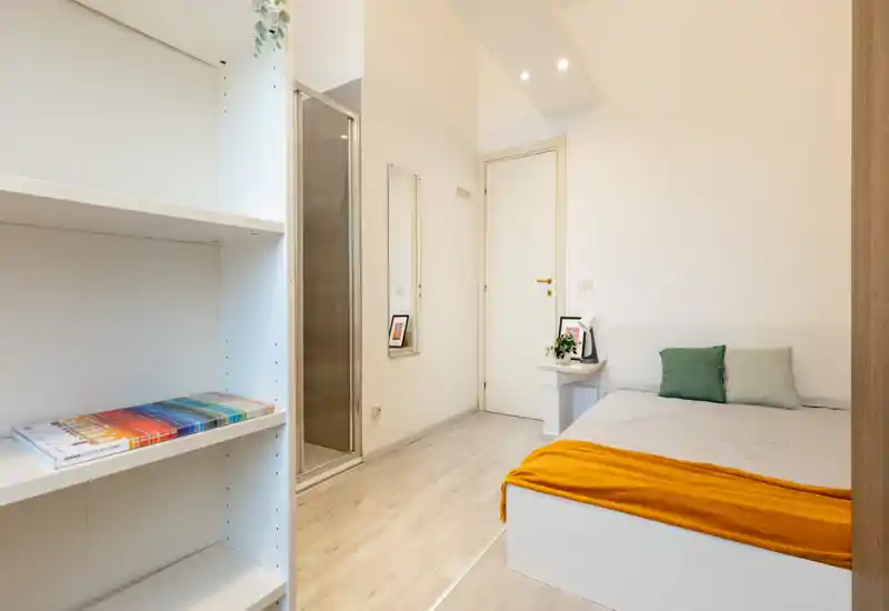Dormitorio individual ordenado con cama baja, manta colorida y cojines; estilo luminoso y minimalista con una mesita y planta que aportan calidez.