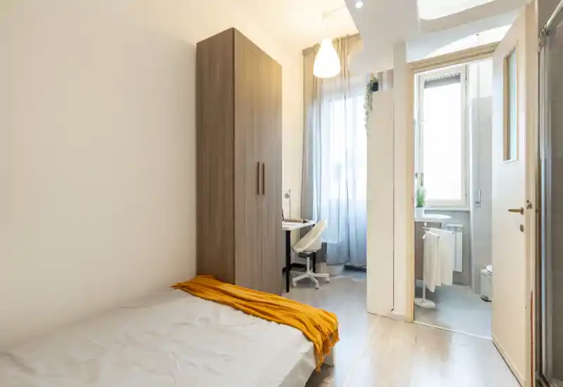 Dormitorio compacto y luminoso con cama individual, armario de madera y pequeño escritorio junto a la ventana; estilo moderno y minimalista con luz natural.