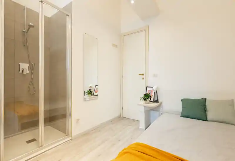 Dormitorio minimalista y luminoso con cama doble, cojines y una manta acogedora; la habitación dispone de una repisa como mesita y un baño con ducha y puerta de cristal.