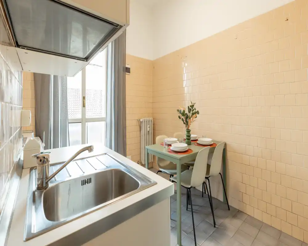 Cocina compacta y luminosa con fregadero de acero inoxidable, mesa pequeña para cuatro y una gran ventana que aporta luz natural.