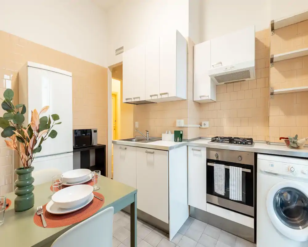 Cocina compacta y luminosa con electrodomésticos modernos, placa de gas y horno integrado. Mesa puesta, nevera, microondas y lavadora hacen el espacio práctico para alquileres de corta estancia.