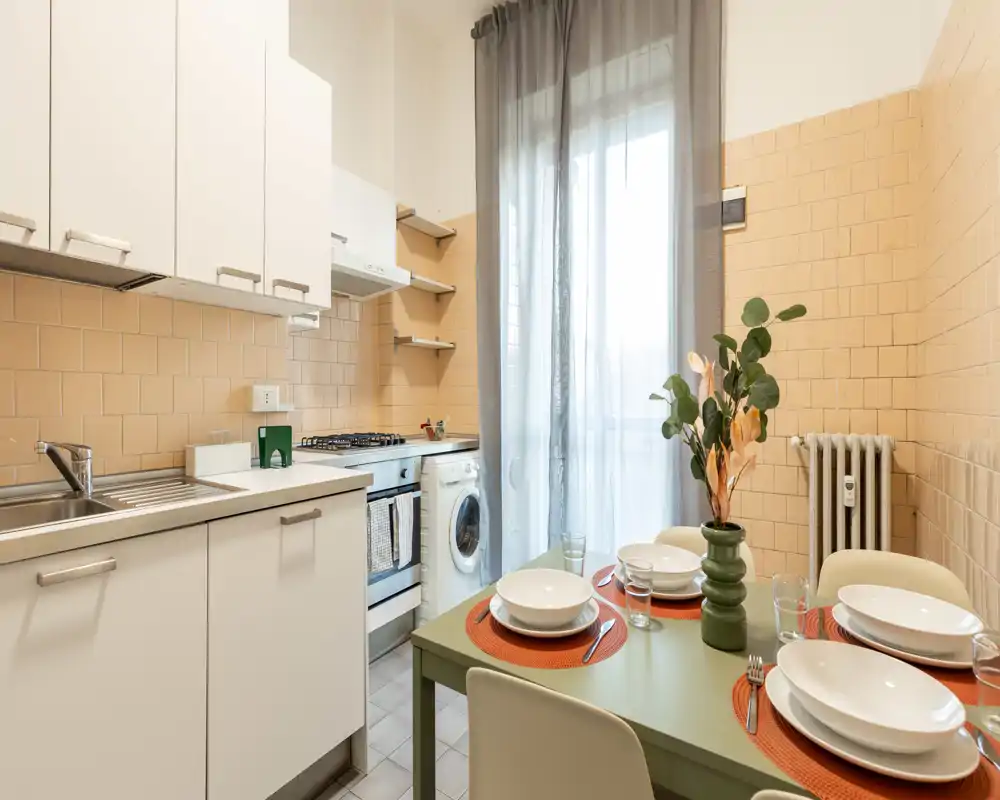 Cocina compacta y luminosa con muebles blancos modernos, cocina de gas y una pequeña mesa puesta para cuatro. La luz natural de la ventana/puerta y las baldosas cálidas crean un ambiente acogedor.