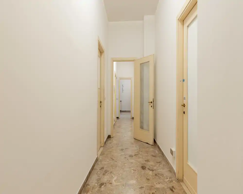Pasillo largo y bien iluminado con varias puertas que dan a otras estancias; paredes neutras y suelo de piedra crean un espacio de entrada/pasillo limpio y minimalista.