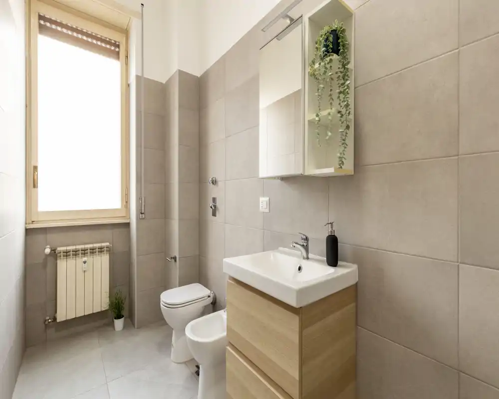 Baño moderno y luminoso con armario-espejo, lavabo con mueble de madera, bidé y váter; azulejos grises neutros y una ventana grande que aporta luz natural.