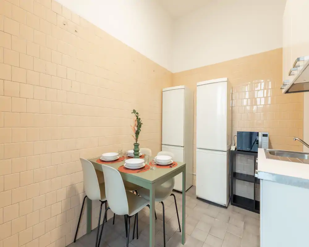 Cocina compacta con una pequeña mesa para cuatro, dos frigoríficos independientes, microondas y fregadero — espacio luminoso y ordenado ideal para comidas informales.