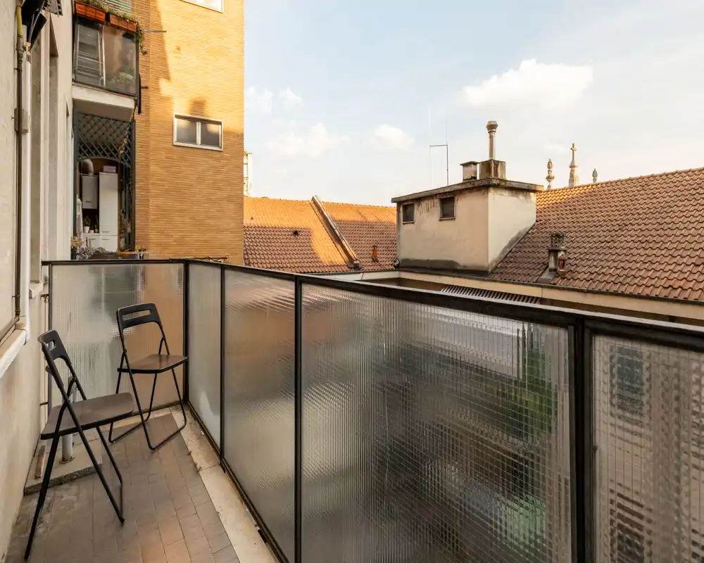 Balcón estrecho y soleado con dos sillas plegables y barandilla de vidrio semiopaco, con vistas a los tejados — perfecto para el café de la mañana.