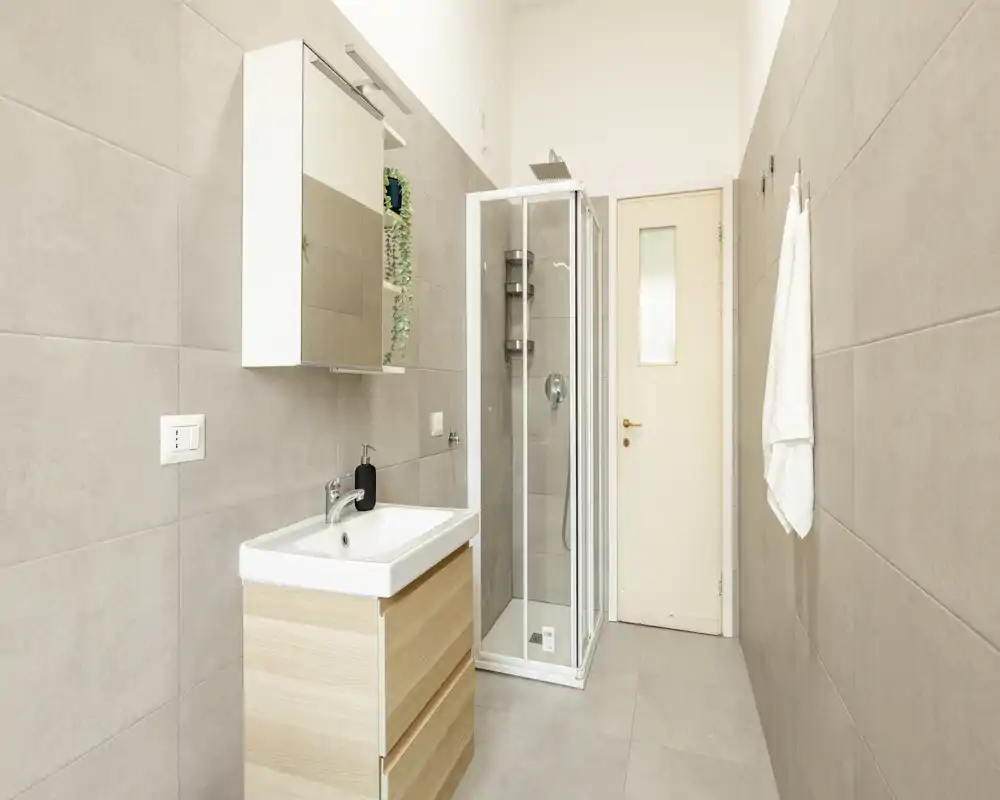 Baño moderno y luminoso con ducha de esquina con mampara de cristal, mueble de lavabo con espejo y azulejos en tonos neutros en paredes y suelo.