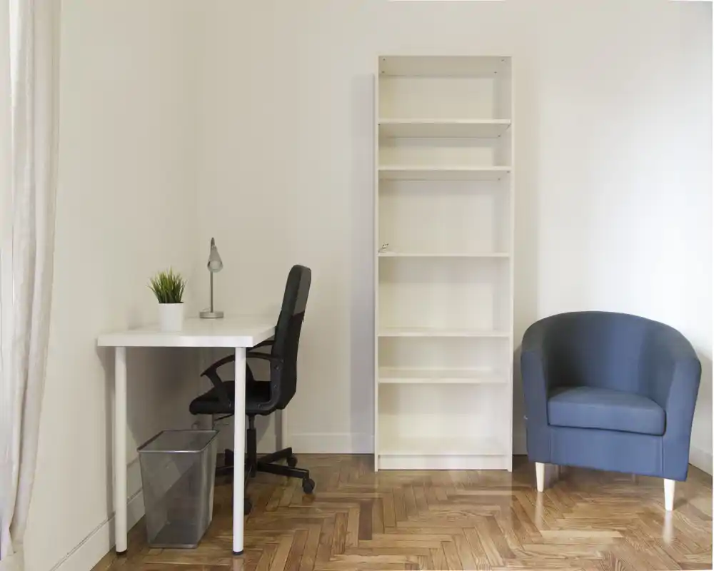 Zona de estar minimal y luminosa con escritorio compacto, estantería y un cómodo sillón azul — apta para trabajar y relajarse.