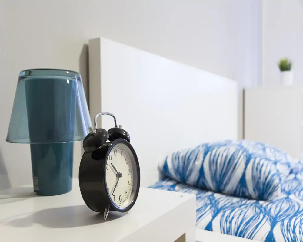 Dormitorio moderno y luminoso en primer plano con mesita de noche, un despertador retro, una lámpara turquesa y cama con ropa de cama azul estampada.