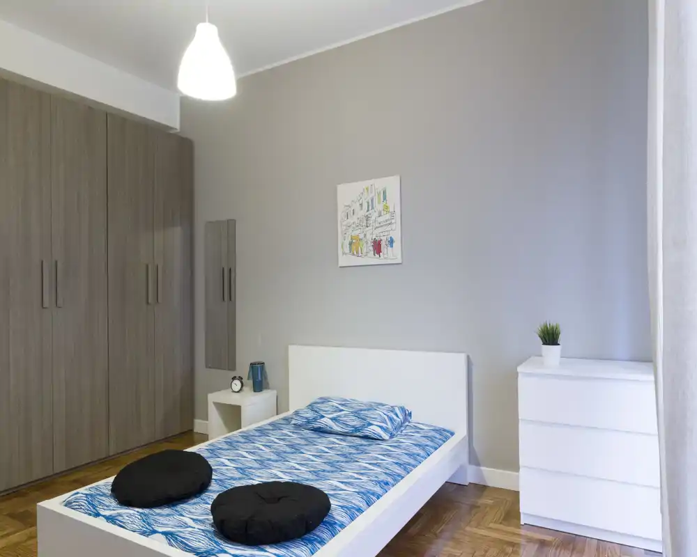 Dormitorio moderno y luminoso con cama blanca baja, ropa de cama azul estampada y mobiliario minimalista. Armario grande, mesita y cómoda con planta, ideal para un anuncio de alquiler.