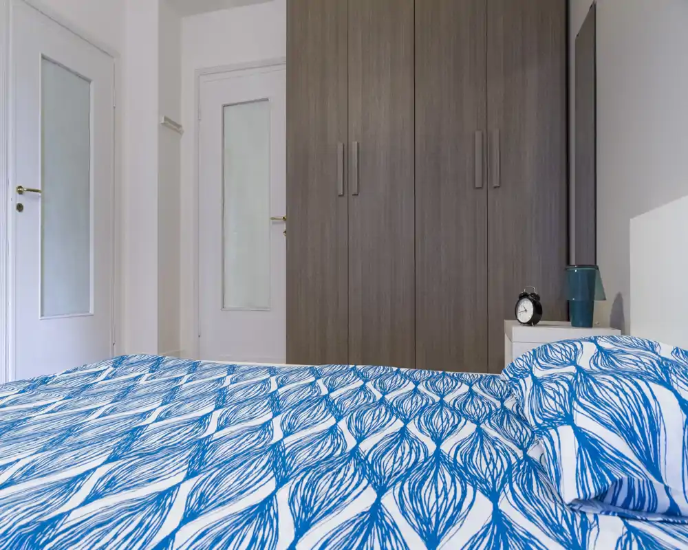 Dormitorio luminoso y de estilo sencillo con un edredón azul estampado en primer plano, un gran armario y una mesita de noche con lámpara y despertador.