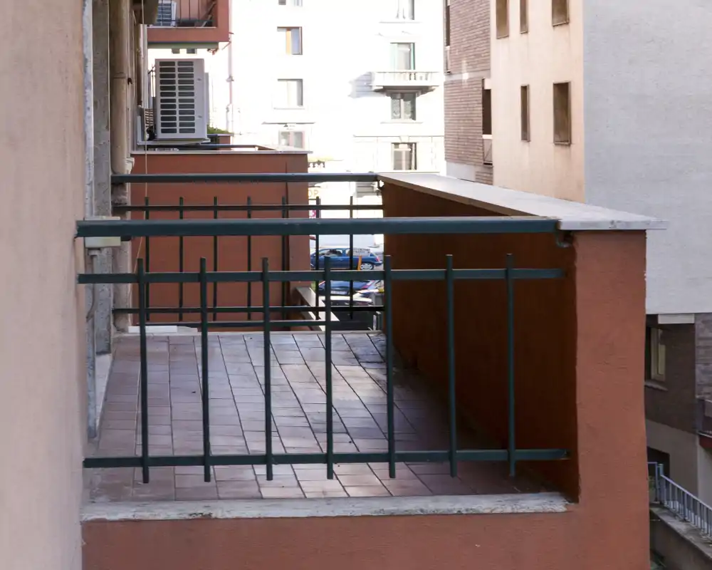 Pequeño balcón urbano con suelo de baldosas y barandilla metálica, con vistas a edificios cercanos. Espacio exterior funcional, apto para una silla o una mesa pequeña.