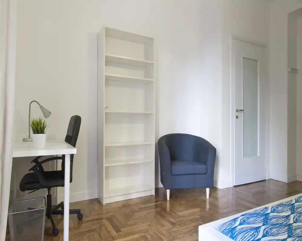 Rincón de dormitorio luminoso y minimalista con esquina de la cama visible, estantería blanca, escritorio y butaca — limpio, moderno y bien iluminado.