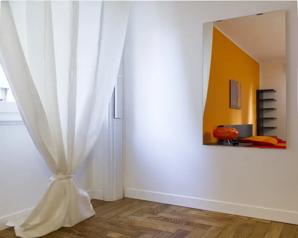Rincón luminoso con una cortina blanca recogida y un espejo en la pared que refleja un dormitorio con pared naranja y una cama. La foto destaca la luz natural y el suelo de parqué.