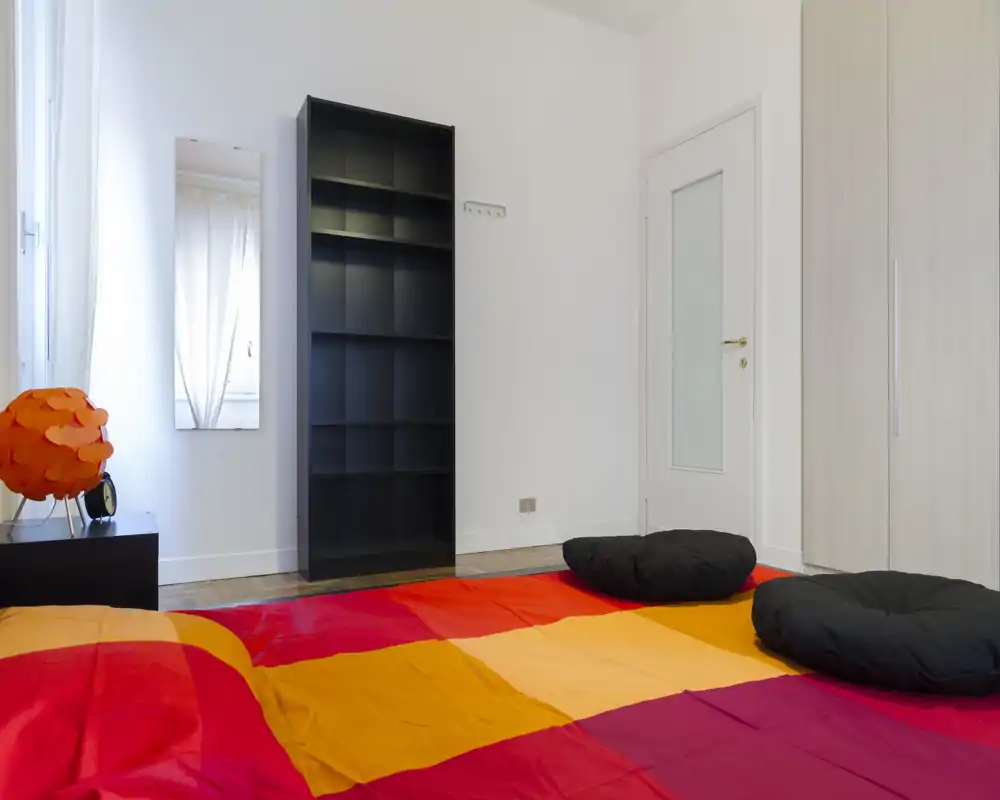 Dormitorio moderno y luminoso con edredón colorido, cojines bajos y almacenamiento sencillo; líneas limpias y luz natural crean un ambiente acogedor.