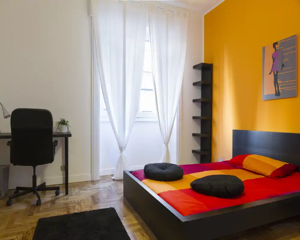 Dormitorio moderno y luminoso con cama baja tipo plataforma, ropa de cama en tonos rojo-naranja y cojines redondos. La habitación cuenta con una gran ventana con cortinas translúcidas, un escritorio con silla y suelo de parqué cálido.
